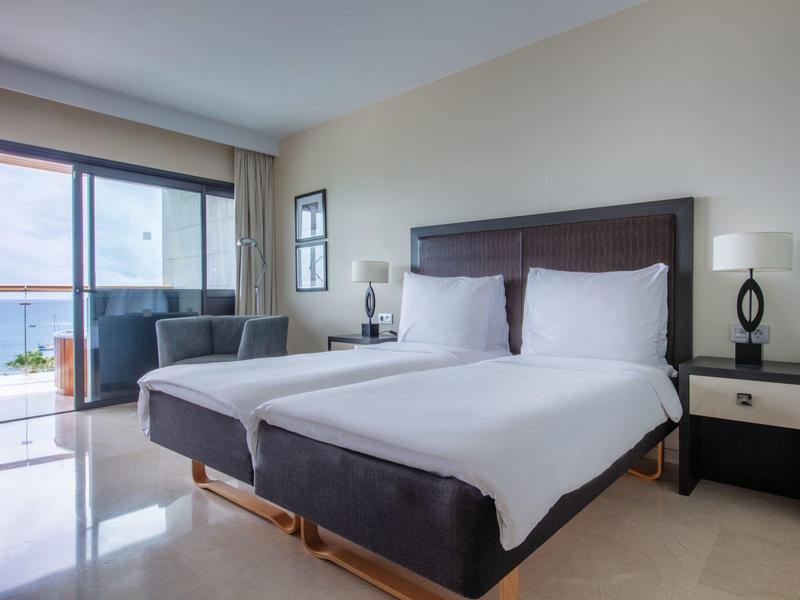 Světlý hotelový pokoj se dvěma jednolůžky a výhledem na moře z balkonu.