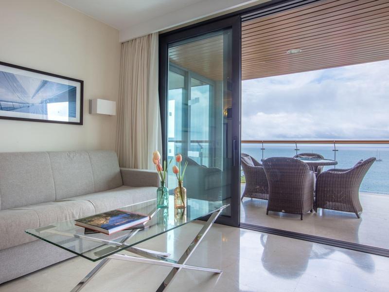 Moderní obývací prostor se skleněným konferenčním stolkem, pohovkou a balkonem s výhledem na moře.