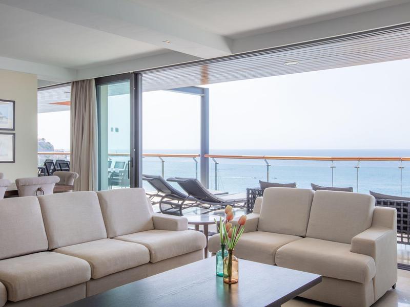 Světlý salón s béžovými pohovkami a výhledem na balkon s lehátky a mořem.