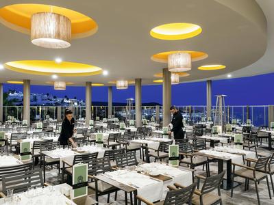 Open hotelrestaurant met moderne lampen en uitzicht op de stad bij schemering.