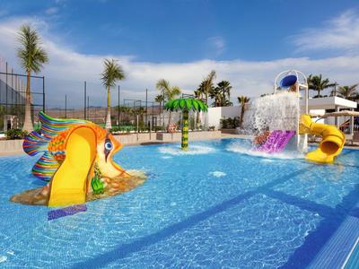 Área de juegos acuáticos colorida con toboganes pequeños y fuentes en la piscina exterior.