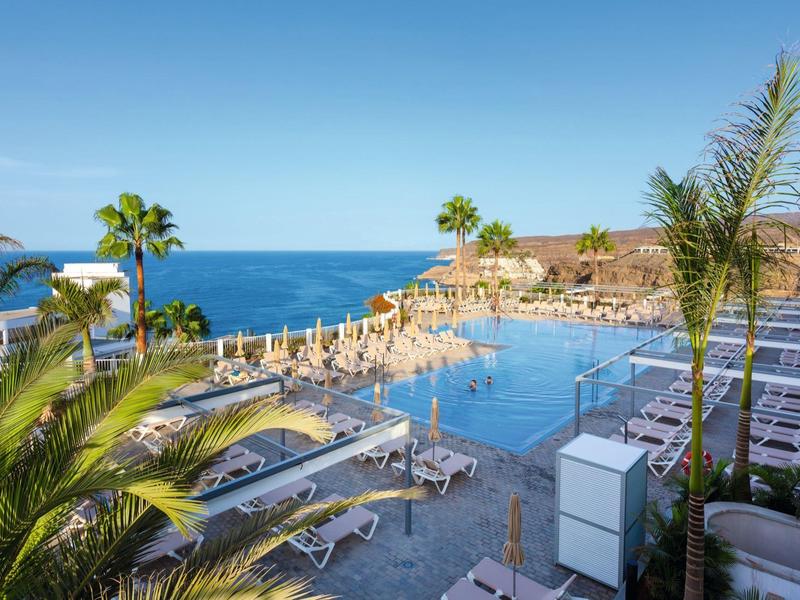 Gran piscina del hotel con tumbonas y vista al mar bajo un cielo azul.