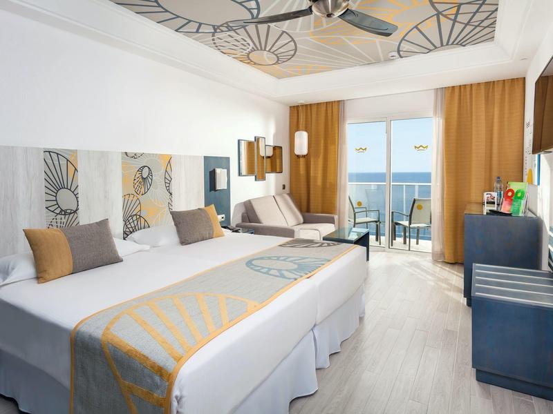 Habitación de hotel luminosa con dos camas, balcón y vista al mar.