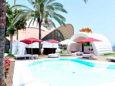 Basen na zewnątrz z leżakami i czerwonymi parasolami przy nowoczesnym hotelu z palmami.