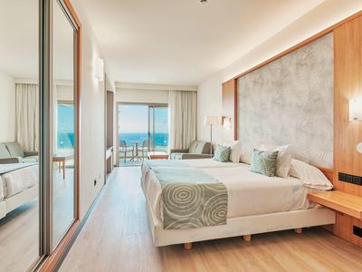 Jasny pokój hotelowy z podwójnym łóżkiem, drewnianymi akcentami i balkonem z widokiem na ocean.