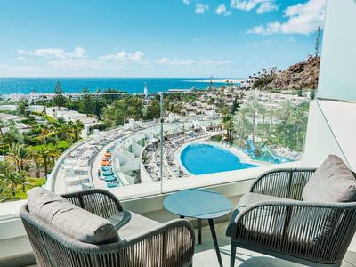 Balkon z krzesłami i stołem z widokiem na basen, ogrody i morze pod niebieskim niebem.