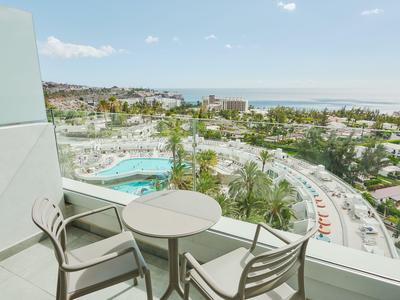 Balcón con mesa y dos sillas con vista a la piscina, jardín y carretera en zona hotelera.