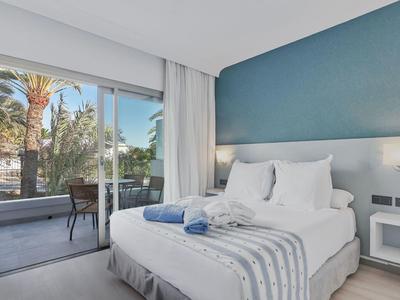 Jasny pokój hotelowy z dużym łóżkiem, niebieską ścianą, balkonem z krzesłami i palmami na zewnątrz.