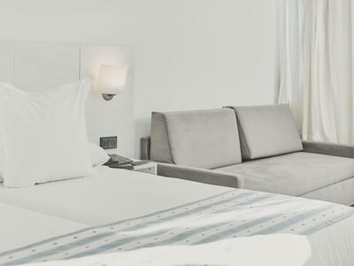 Jasny pokój hotelowy z białym łóżkiem i jasnoszarym sofą przy oknie z zasłonami.