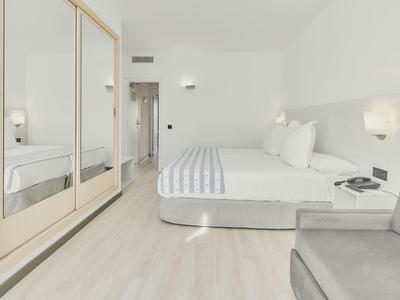 Jasny pokój hotelowy z dużym łóżkiem, pasiastą pościelą, lustrowaną szafą i szarą sofą.