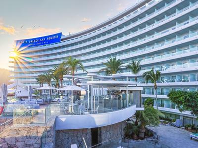 Hotel moderno con edificio de vidrio curvado, palmeras y sombrillas al atardecer.