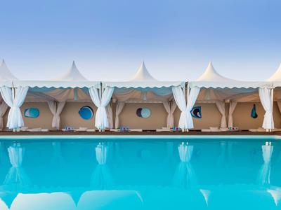 Blauer Pool vor weißen Zelten mit Vorhängen und runden Fenstern unter klarem Himmel.