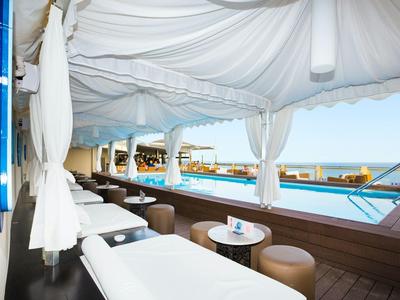 Strefa wypoczynkowa przy basenie z białymi leżakami, zasłonami i widokiem na ocean.