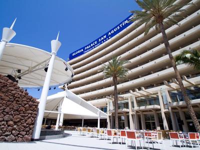 Duży hotel z zakrzywionymi balkonami, palmami i miejscami do siedzenia na zewnątrz pod białymi markizami.