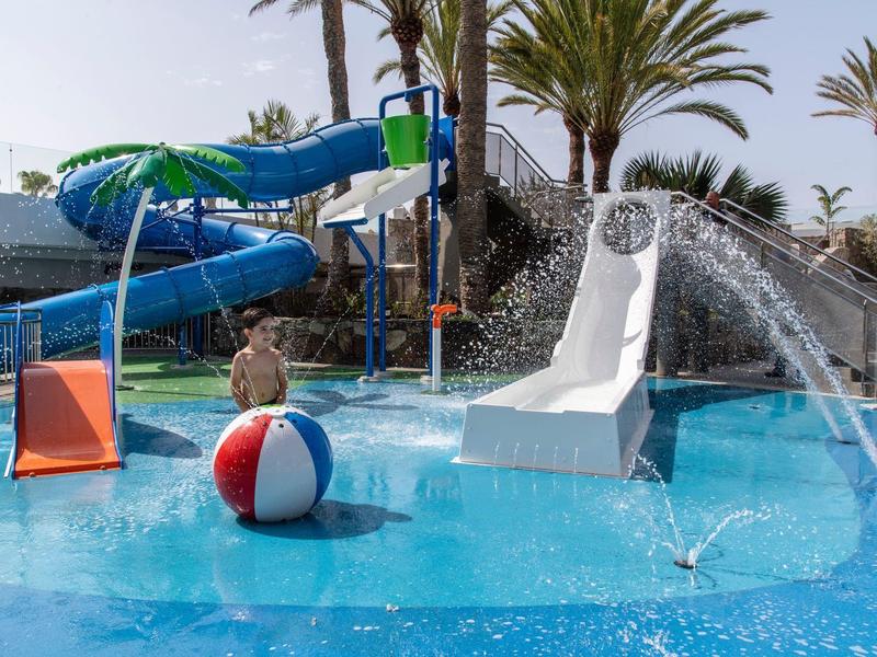 Dzieci bawią się w basenie z zjeżdżalniami, fontannami i dużą piłką plażową pod palmami.