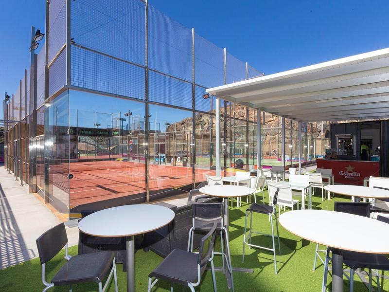 Taras na zewnątrz z białymi stołami i krzesłami obok kortu tenisowego pod czystym, niebieskim niebem.