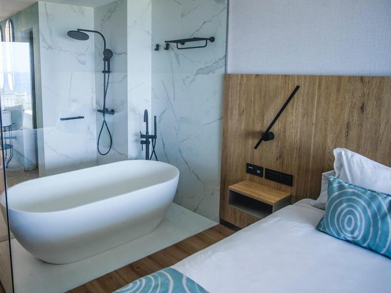 Nowoczesny pokój hotelowy z łóżkiem i przestronną łazienką z wolnostojącą wanną i szklaną kabiną prysznicową.