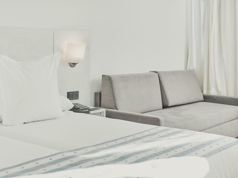 Jasny pokój hotelowy z białym łóżkiem i jasnoszarym sofą przy oknie z zasłonami.