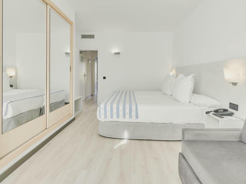 Jasny pokój hotelowy z dużym łóżkiem, pasiastą pościelą, lustrowaną szafą i szarą sofą.