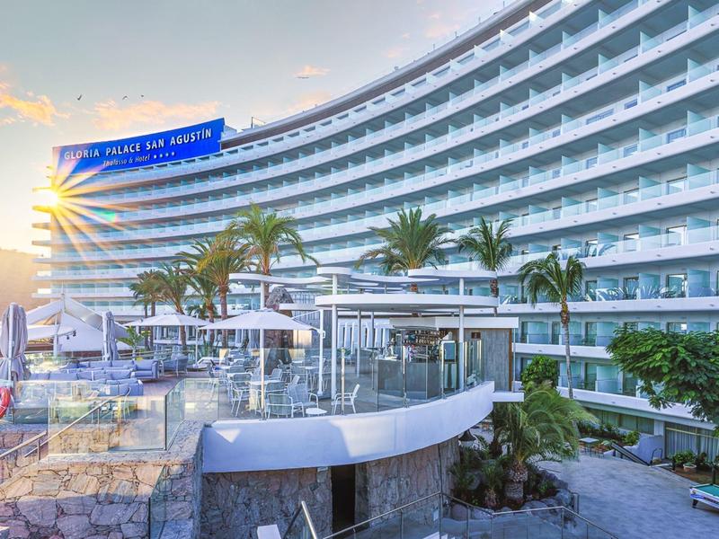 Zakład hotelowy o zakrzywionym kształcie z balkonami, palmami i basenem o zachodzie słońca.