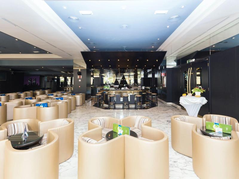 Nowoczesny hotelowy salon z beżowymi fotelami wokół małych stolików i centralnym barem.