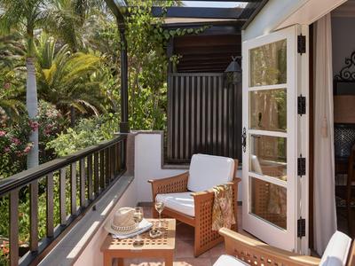 Gemütlicher Balkon mit Korbsessel, Kissen, Holztisch und Glasgeschirr, umgeben von grüner Pflanzenwelt.