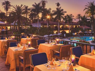 Outdoor dining on a restaurant terrace by a lit pool and palm trees at dusk.