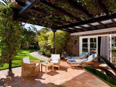 Gemütliche Terrasse mit Holzdach, Gartenmöbeln aus Holz und weißen Kissen, umgeben von grünem Garten.