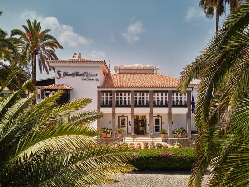 Großes weißes Gebäude mit roten Ziegeldach, umgeben von Palmen und grünem Garten.