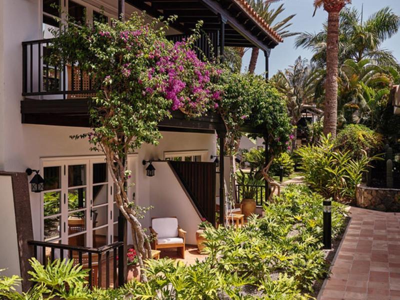 Zweistöckiges Haus mit Balkon, umgeben von Palmen, blühenden Pflanzen und gepflastertem Weg.