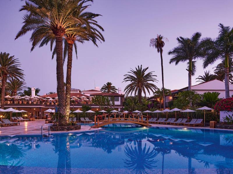 Großer Pool mit Palmen und Sonnenliegen bei Abenddämmerung in tropischer Hotelanlage.
