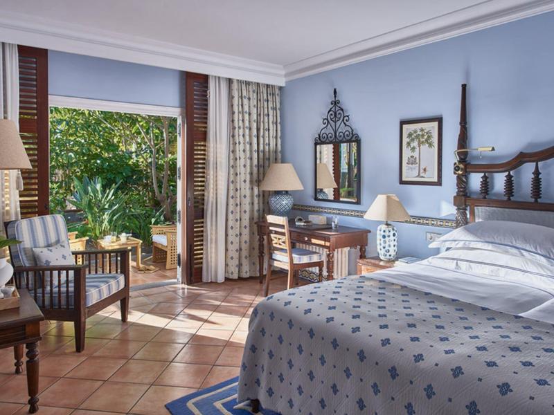 Helles Schlafzimmer mit Holzmöbeln, blauem Bett, zwei Sesseln und Terrasse mit Gartenblick.