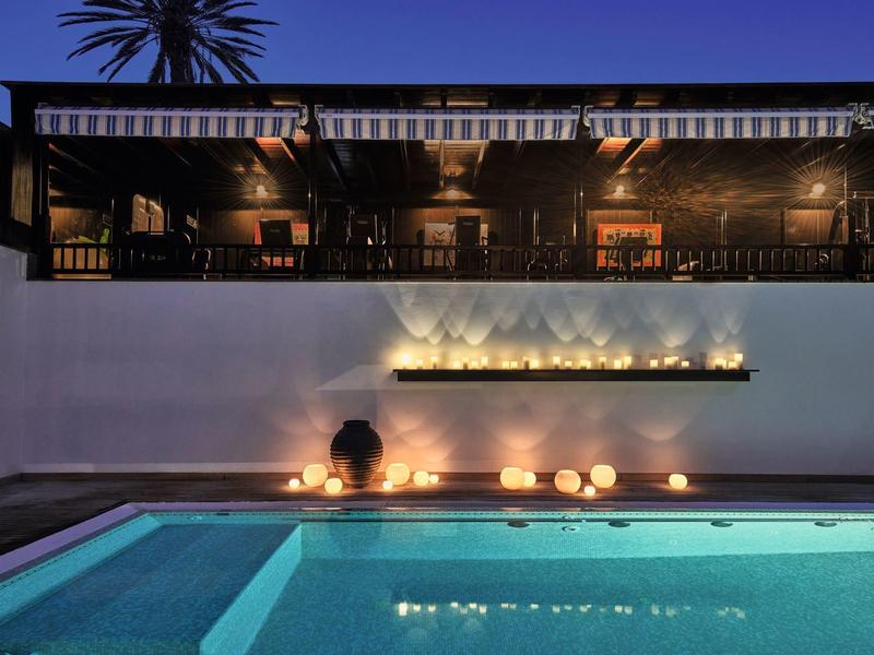 Beleuchteter Pool vor modernem Haus mit Dachterrasse und Pflanzen bei Nacht.