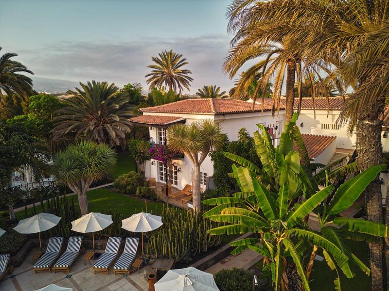 Ein weißes Haus mit roten Dachziegeln, umgeben von grünen Palmen und Sonnenschirmen im Garten.