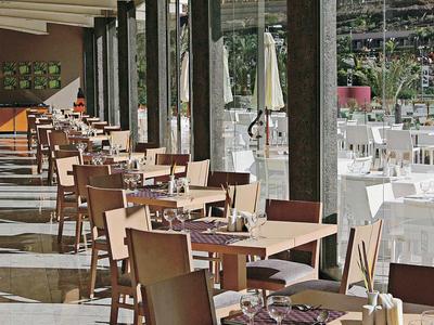Lange terras met houten tafels en beige stoelen, omgeven door glazen wanden en een plafond met houten balken.
