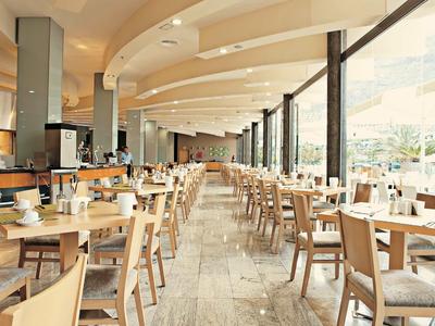 Lichte eetzaal met houten tafels, stoelen en grote ramen in een vakantiehotel.