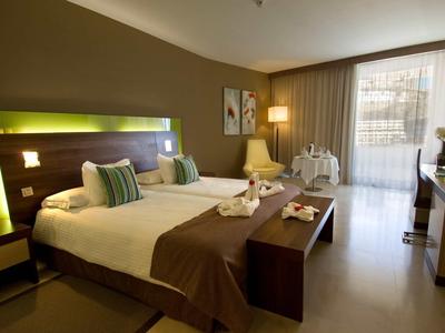 Modern hotelkamer met groot bed, bankje, verlichting en balkon met gordijnen.
