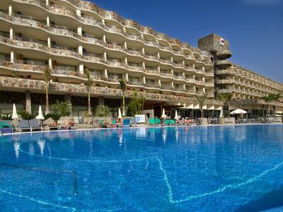 Large hotel with multiple floors, balcony rooms, and a large blue pool in the foreground.