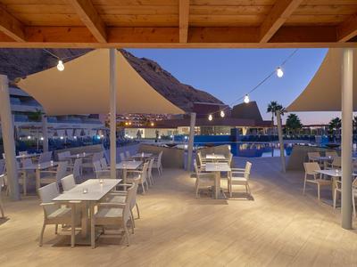 Restaurante al aire libre con mesas y sillas blancas bajo un techo de madera al atardecer junto a un lago de montaña.