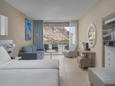 Habitación de hotel moderna con cama doble, área de descanso y balcón con vista a las rocas.