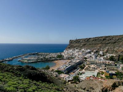 Paisaje costero con cielo azul claro, mar y un pequeño puerto frente a una montaña rocosa.