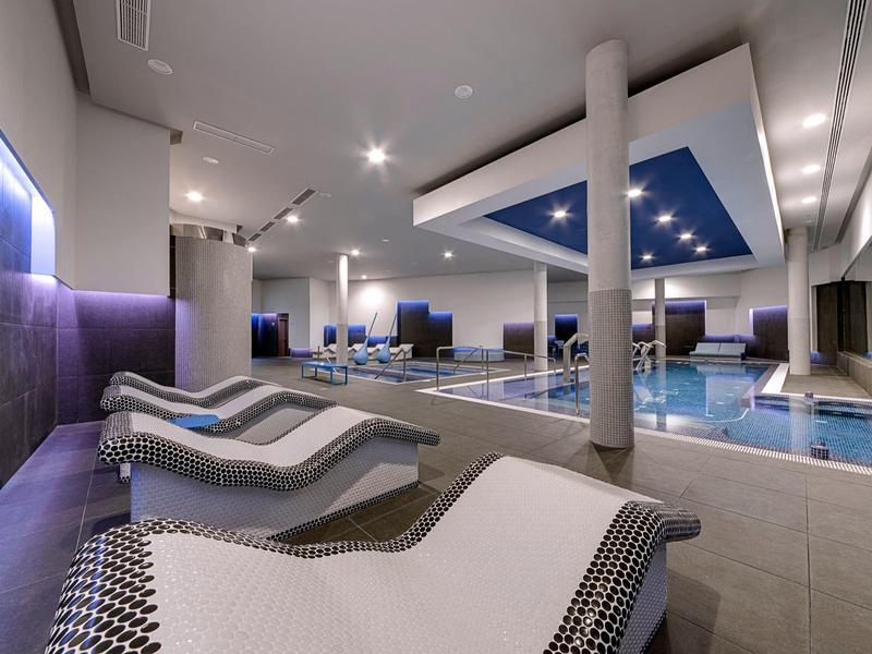 Área moderna de bienestar con piscina climatizada, columnas y tumbonas con iluminación tenue.