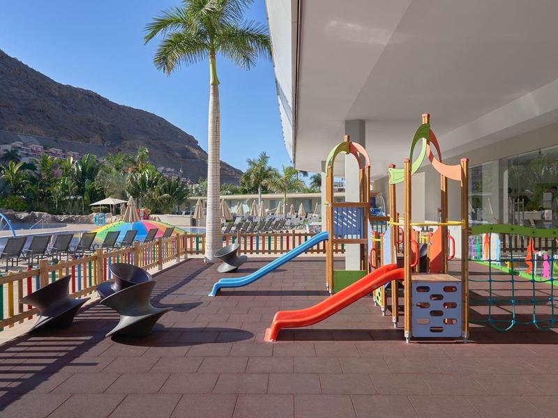 Área de juegos colorida para niños junto a la piscina con montañas y palmeras al fondo.