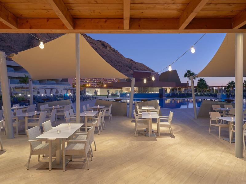 Restaurante al aire libre con mesas y sillas blancas bajo un techo de madera al atardecer junto a un lago de montaña.