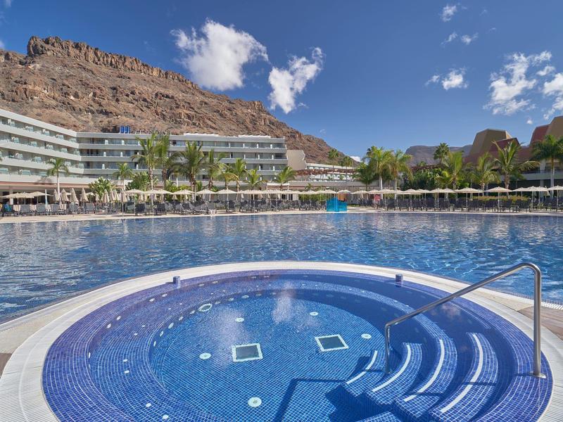 Gran piscina exterior y jacuzzi con vistas a rocas y edificios del hotel bajo cielo azul.