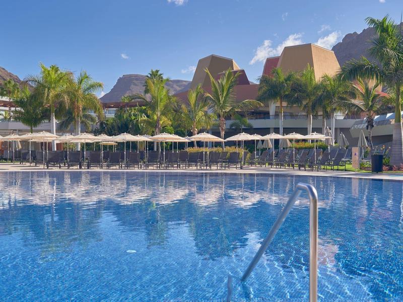 Gran piscina de hotel con tumbonas, sombrillas y palmeras al fondo bajo cielo azul.