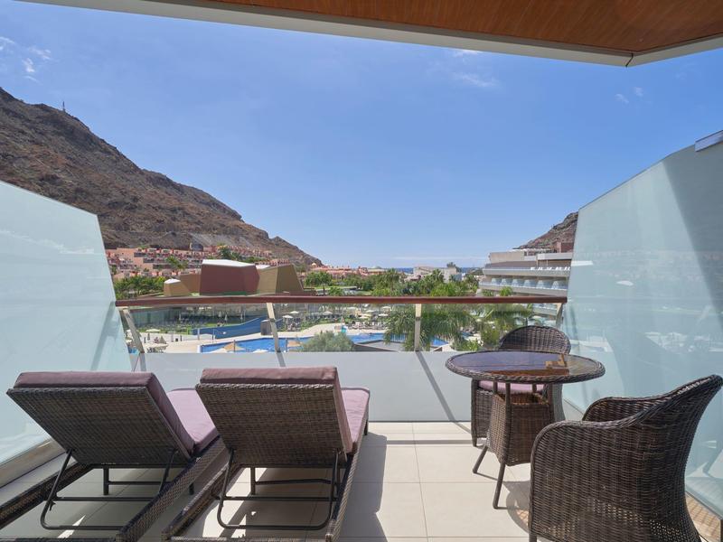 Balcón con dos tumbonas, mesa y silla con vista a una piscina y montañas.