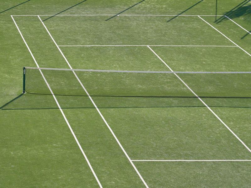 Cancha de tenis verde con líneas blancas, red y sombras bajo el sol.