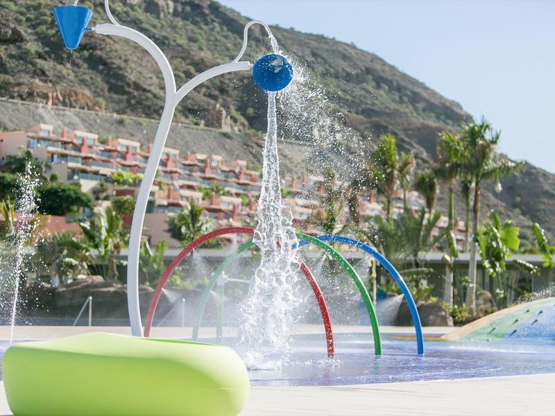 Parque acuático con chorros de agua de colores, palmeras y montañas al fondo bajo cielo despejado.