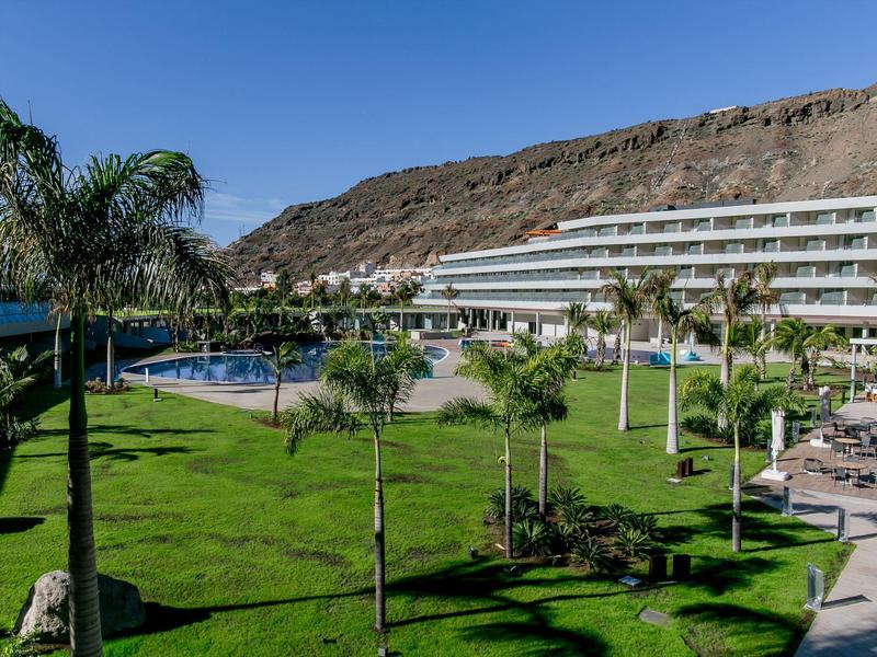 Complejo hotelero con piscina, palmeras y césped frente a una montaña rocosa bajo un cielo despejado.